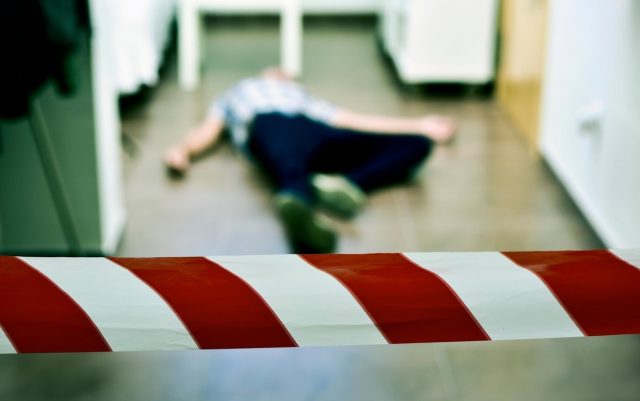 detail of a crime scene with the dead body of a young man lying in the floor indoors, with a red and white police tape in the fo